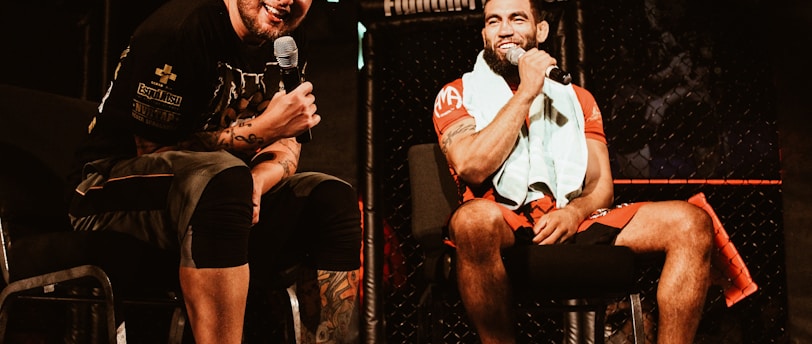 Two individuals are seated on a stage, each holding a microphone and smiling. They are in a sports-related setting with one wearing a casual black outfit and the other in sportswear, with a towel around his neck. The background includes elements of a fighting or martial arts cage.
