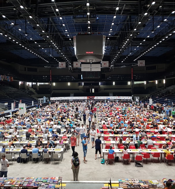An indoor sports arena filled with rows of tables, each occupied by people engaged in competitive activities. The ceiling is high, with numerous lights illuminating the large space. The audience seating on either side is empty, and there are flags hanging from the upper level.