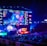 A large esports arena featuring a massive screen displaying a video game, surrounded by sponsor logos and team names. The auditorium is filled with spectators watching the gaming event. Neon blue and purple lights illuminate the stage, creating a vibrant atmosphere.
