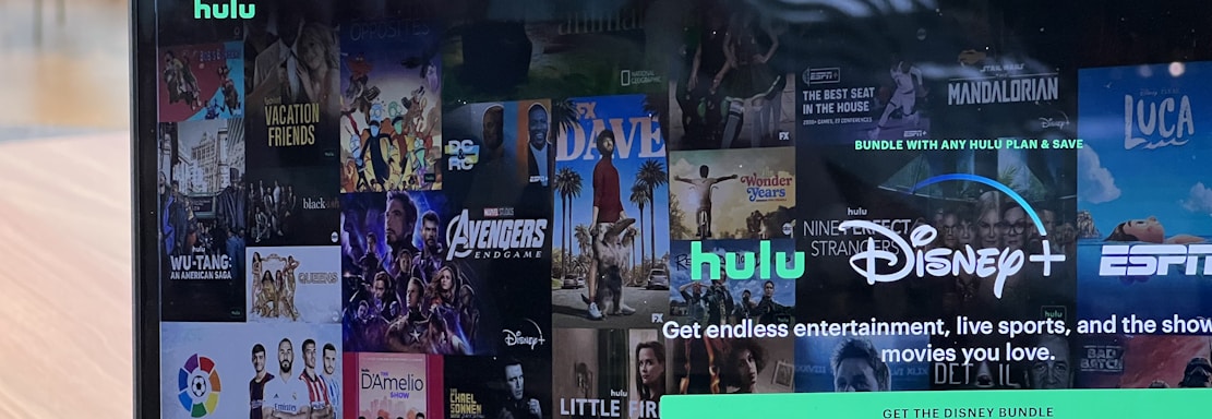 A laptop screen displays a web page featuring a mix of movie and TV show covers, highlighting popular titles and a Hulu and Disney+ bundle promotion. There are visible icons and images of films and shows alongside promotional text offering entertainment and live sports.