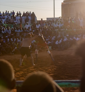 A vibrant wrestling match taking place outdoors, surrounded by a large crowd of spectators. The atmosphere is charged with excitement as people stand and sit around the dirt arena, intently watching the competitors. The sun is setting, casting a warm glow over the scene.