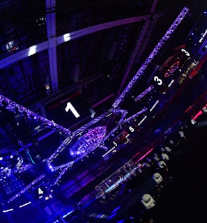 A large indoor arena with colorful and vibrant lighting, featuring a center stage surrounded by rigging and structures. Large numbered screens are visible, indicating marked sections from one to five. Various pieces of equipment and people are scattered around the venue.