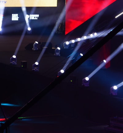 An array of bright stage lights beams across a dark arena, creating a dynamic atmosphere with a large screen displaying various logos and text. A camera crane is visible in the foreground, suggesting a professional event setup.