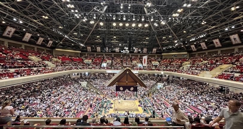 A large indoor arena packed with spectators seated in tiers around a central platform. The platform features traditional Japanese decor, indicating it may be used for sumo wrestling. The atmosphere is bustling, with people filling the seats and many spectators visible in the foreground.