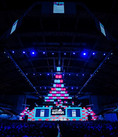 An expansive venue filled with a large audience, featuring a grand stage lit with vibrant blue and pink lights. The ceiling is decorated with a large display, and numerous screens showcase the event's branding. The atmosphere is dynamic and futuristic, reflecting a technologically advanced conference.